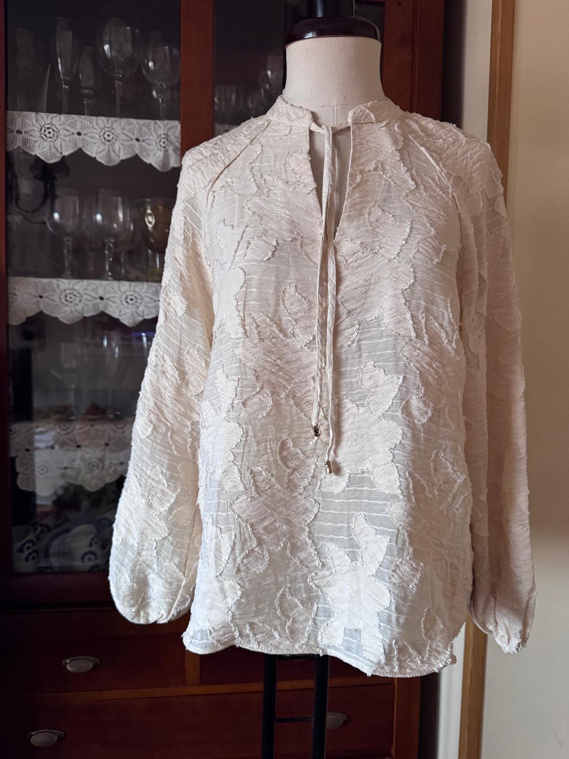 Textured white blouse with a floral design on a mannequin in front of a glass cabinet with glasses and decorative lace.