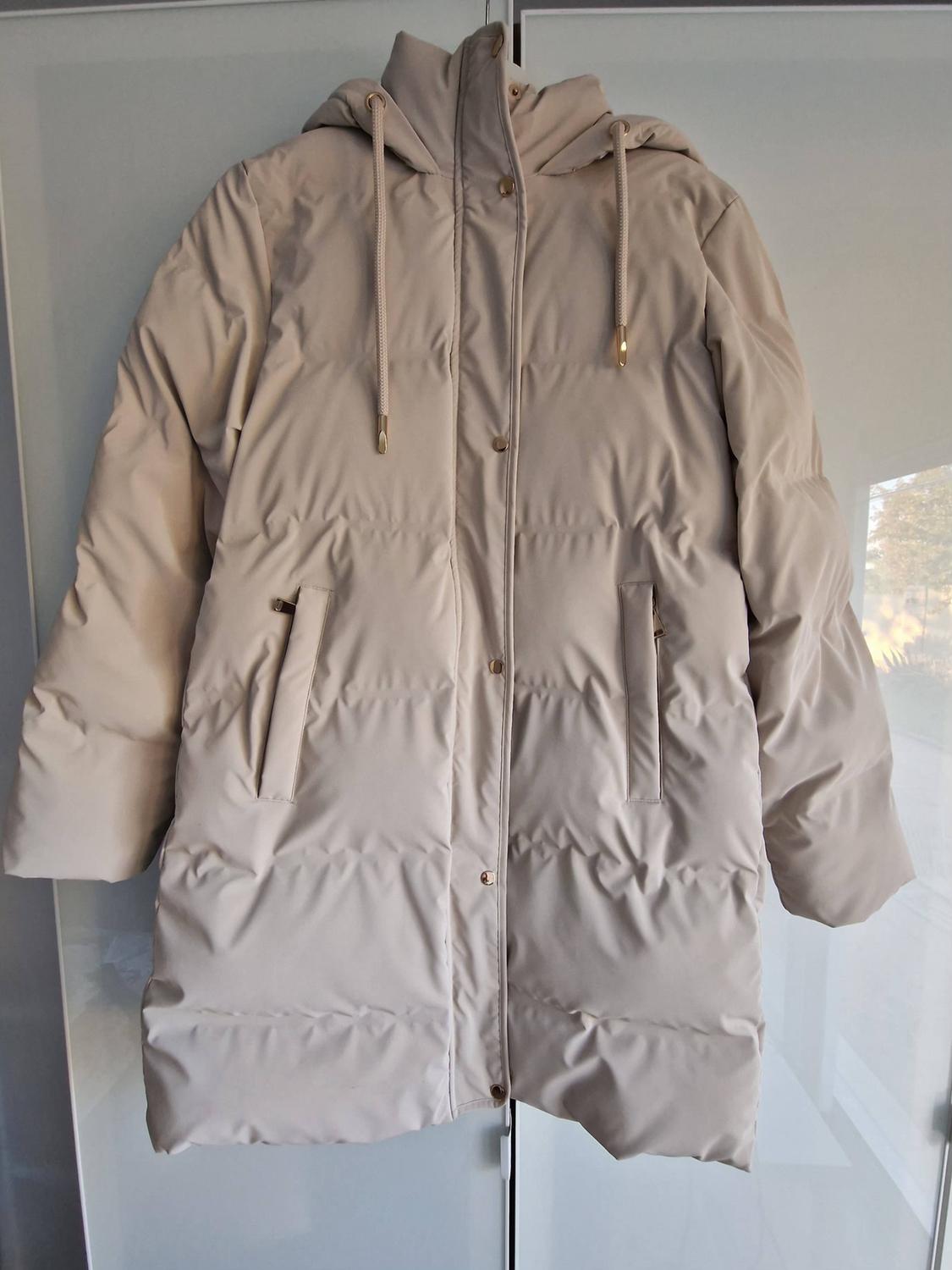 Beige padded coat with a hood, drawstrings and gold-coloured buttons, hanging in a wardrobe with glass doors.