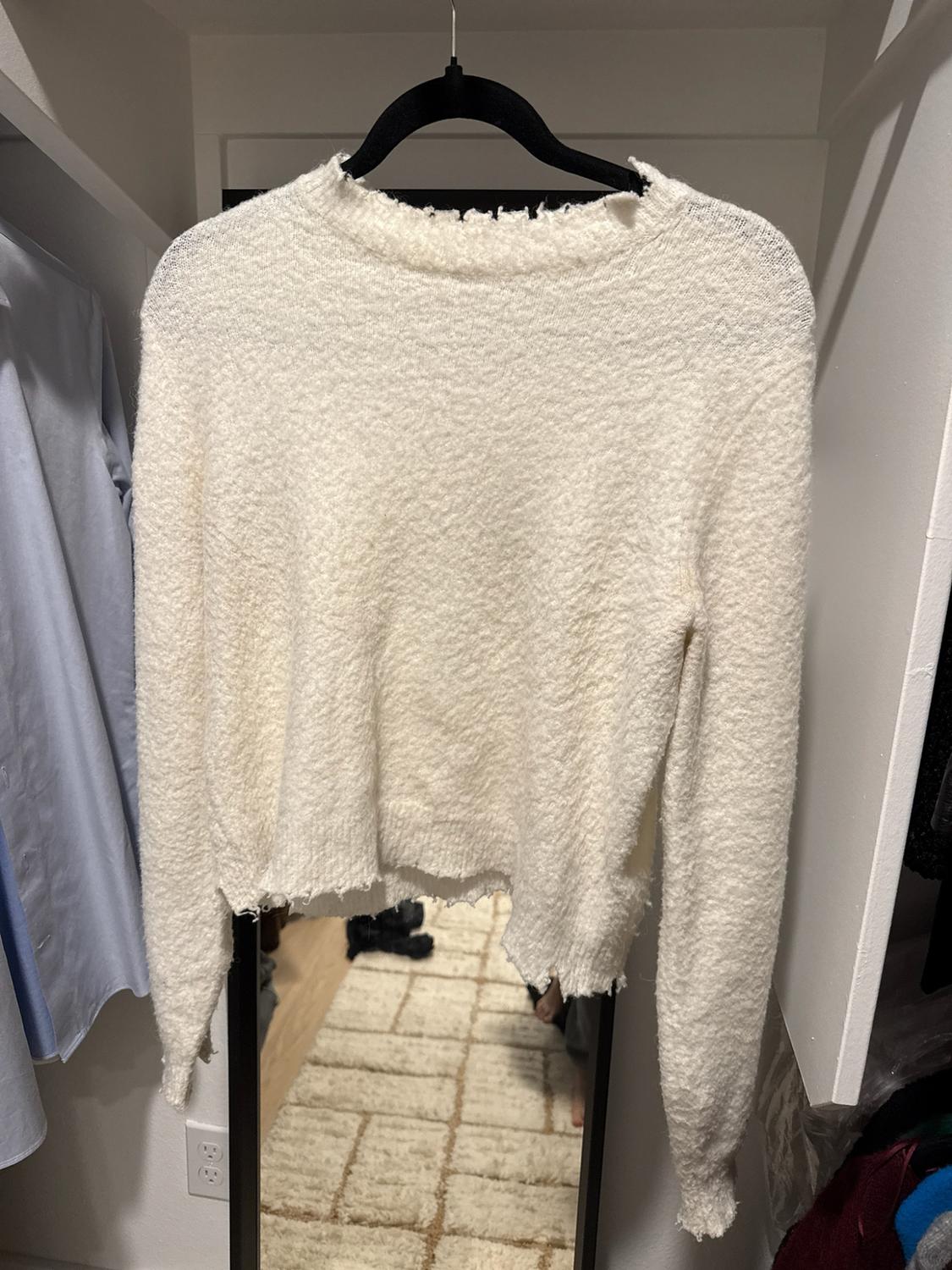 Textured knit white sweater with frayed edges, hanging on a black hanger inside a closet with a mirror in the background.
