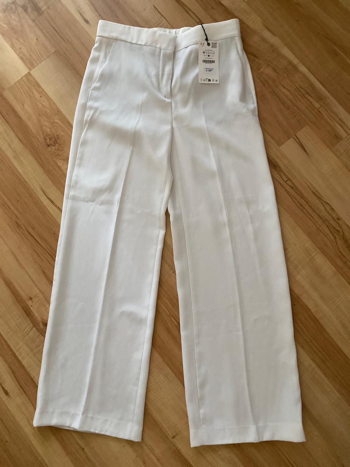 White wide-leg trousers on a light wood floor, with a visible hang tag at the waist.