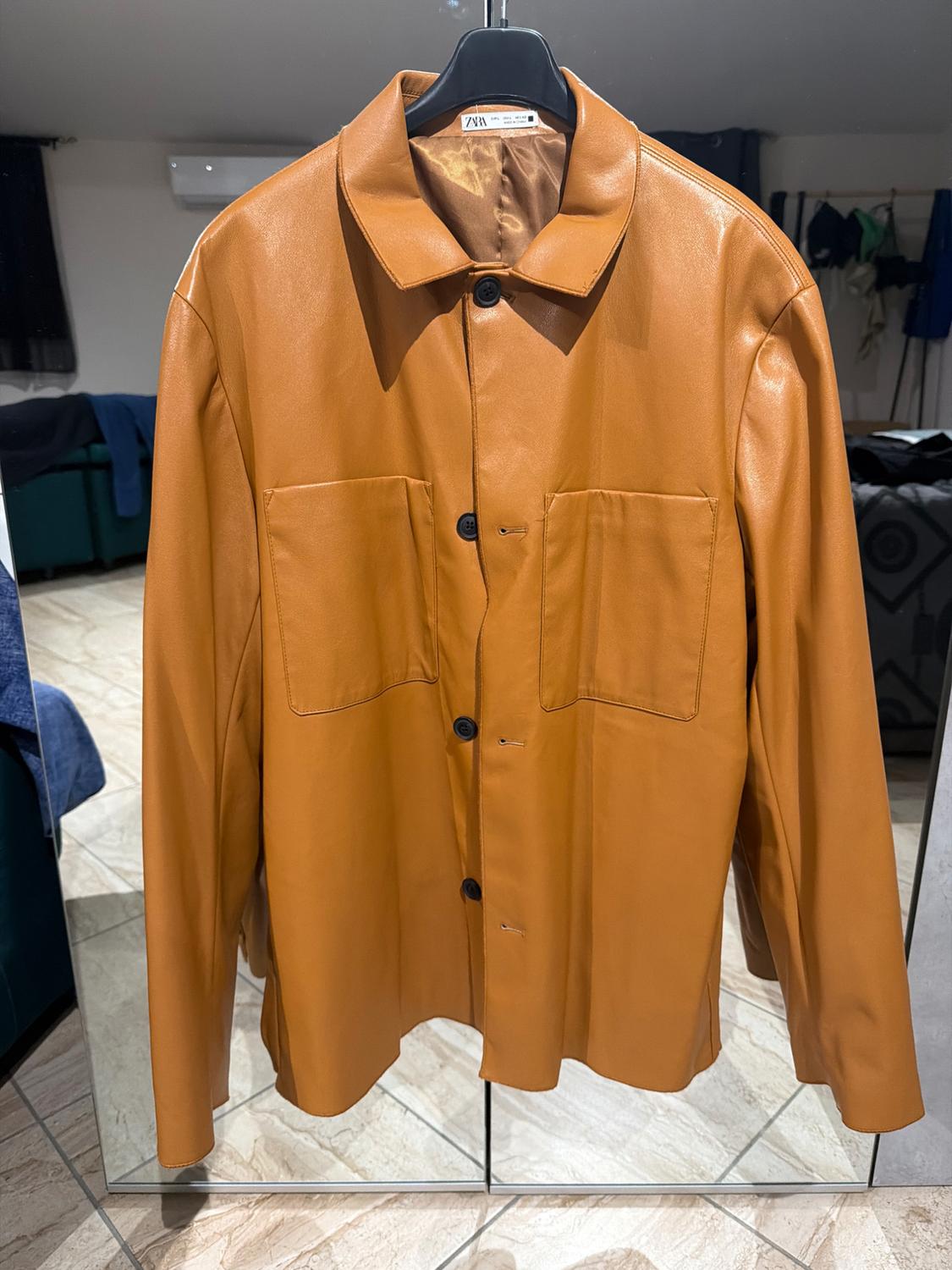 Brown jacket hanging on a black hanger, with dark buttons and large front pockets. Reflection in a mirror in a room.