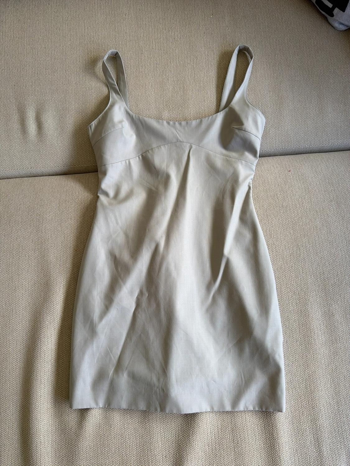 Light beige short dress with wide straps and a round neckline with seams, laid out on a neutral textured fabric surface.