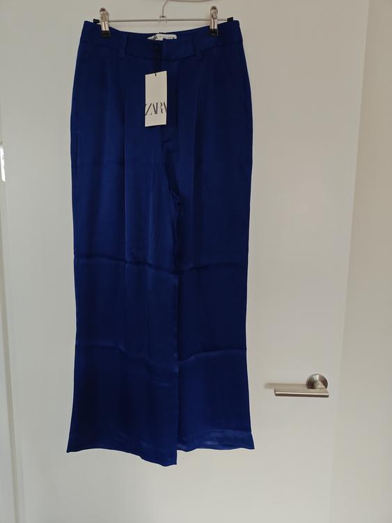 Dark blue trousers hanging on a hanger with a label that says "ZARA", against a white door background.