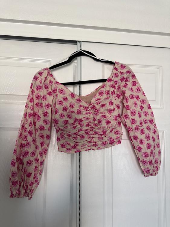 Long-sleeved blouse with pink floral print on a white background, hanging on a black hanger in front of a white door.