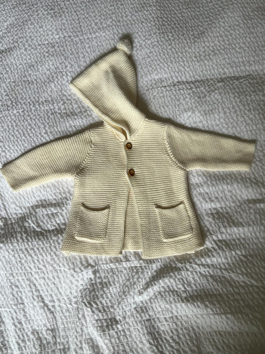 Cream knitted baby cardigan with hood, pom-pom, buttons and pockets, laid out on a textured white blanket. Cosy style.