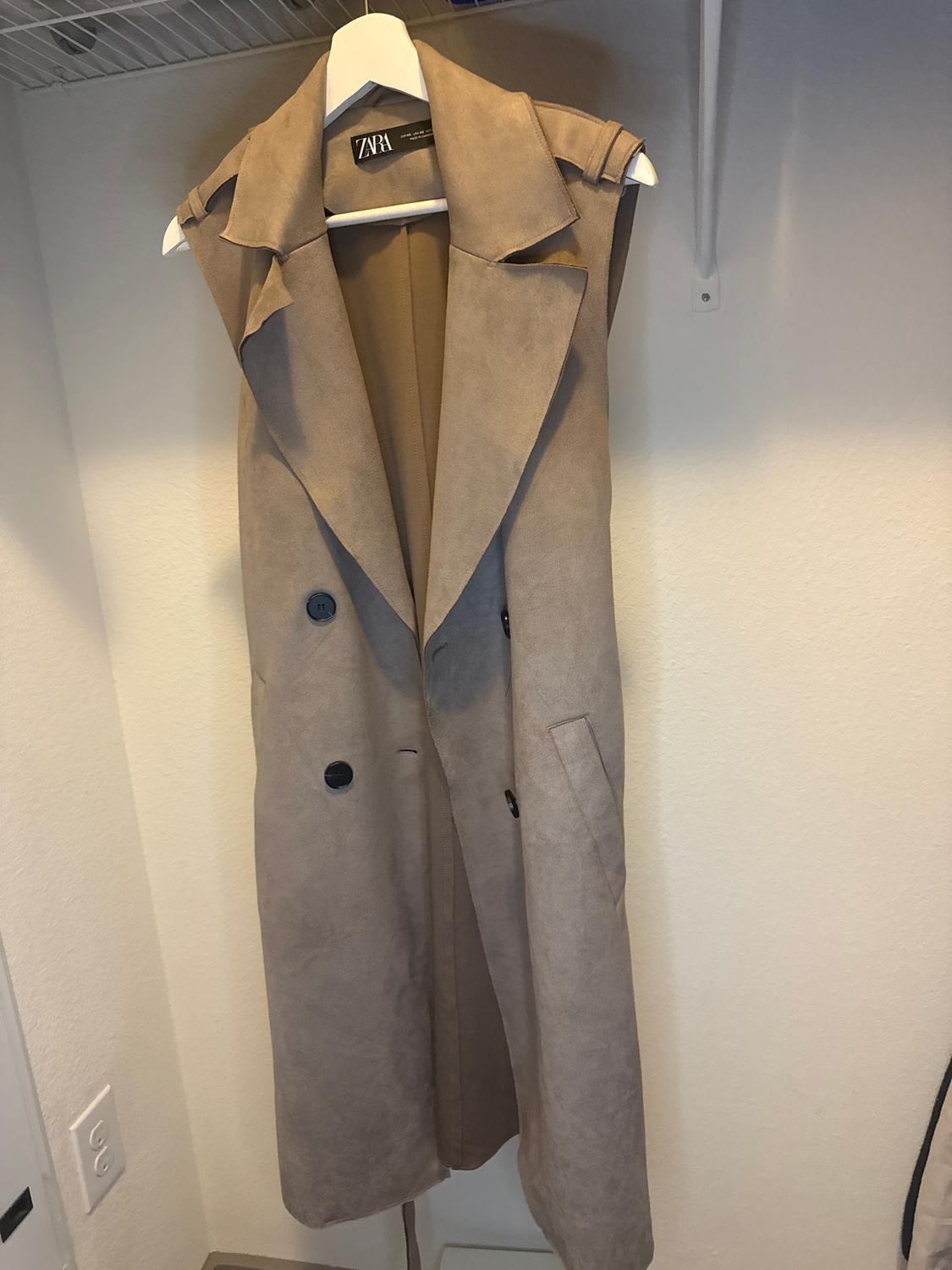 Long beige sleeveless waistcoat hanging on a white hanger, with black buttons and a visible Zara label in a wardrobe.