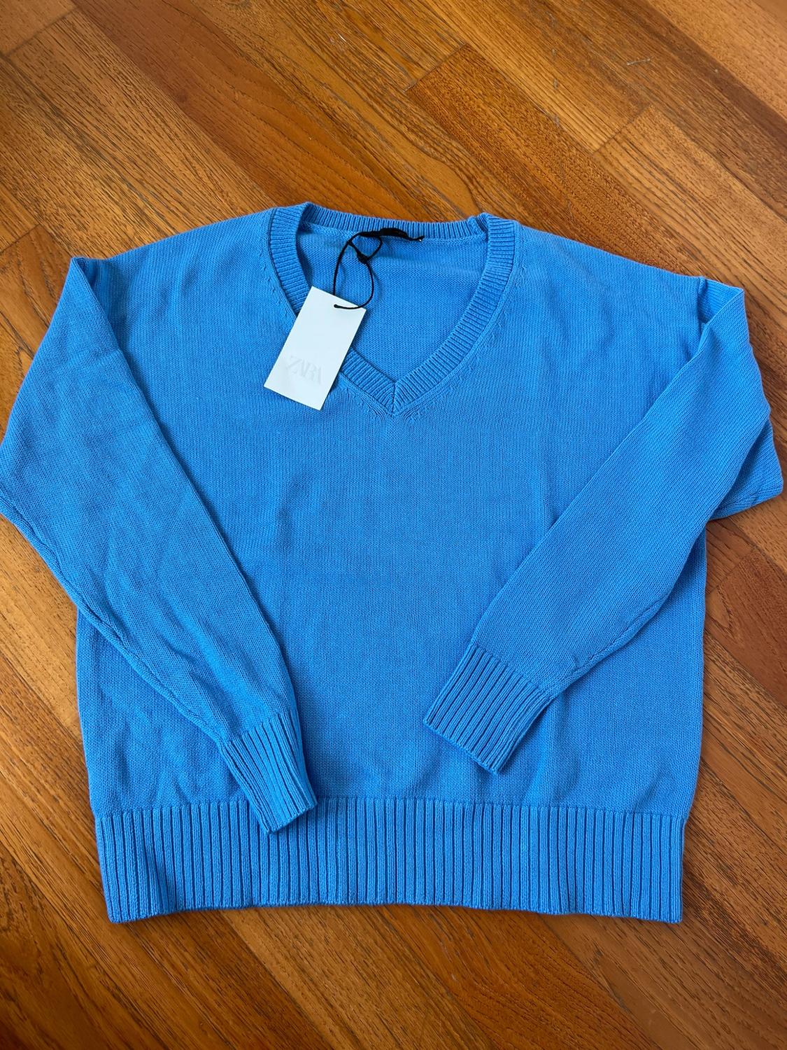 Blue knitted jumper with a V-neck and long sleeves on a wooden floor. It has a white label bearing the text ZARA.
