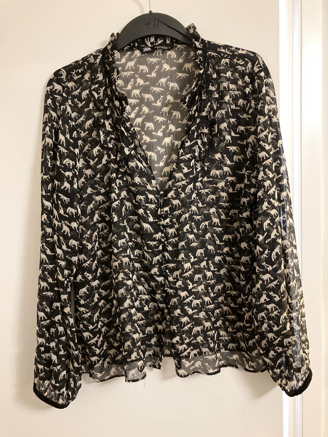 Semi-sheer black blouse with white animal print, long sleeves and ruffles, hanging on a hanger. Text: H&M, ZARA.