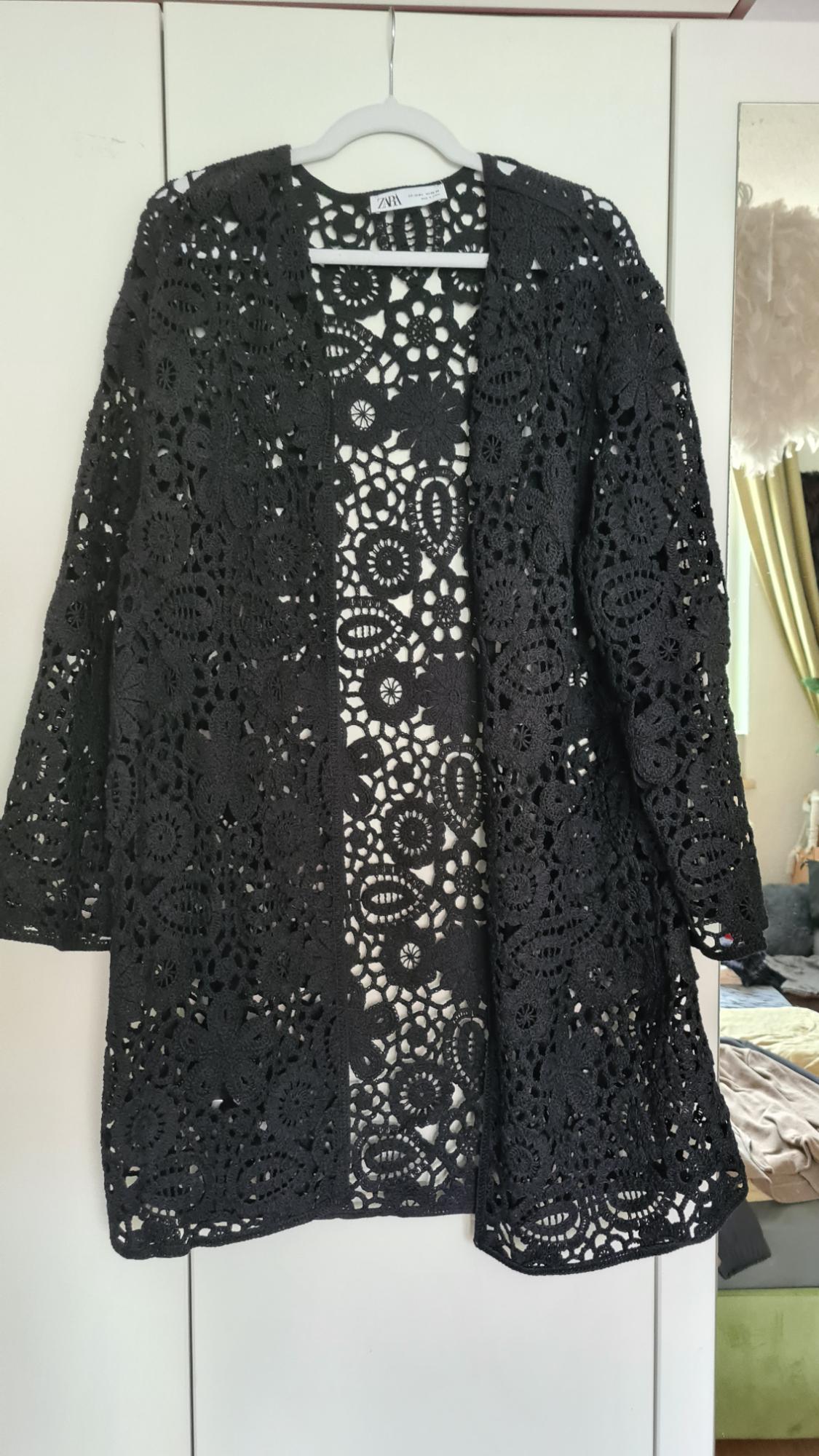 Black lace jacket hanging on a white coat hanger, with floral design and light background. Label visible on the neck.