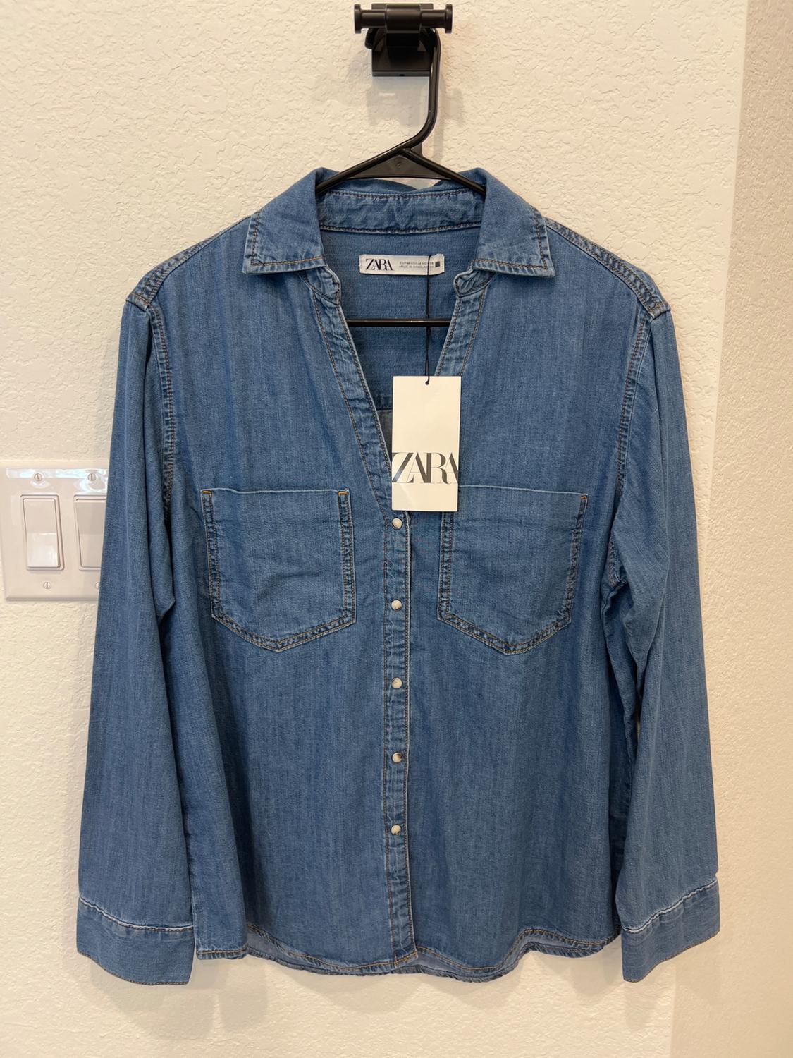 Blue denim shirt with front pockets, hanging on a black hanger. Visible label with the text "ZARA". Light wall background.