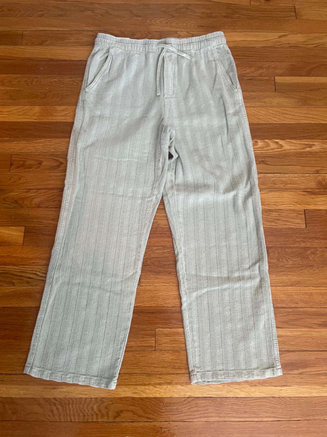 Light grey casual trousers with a vertical stripe texture and an elasticated drawstring waist, laid out on a wooden floor.