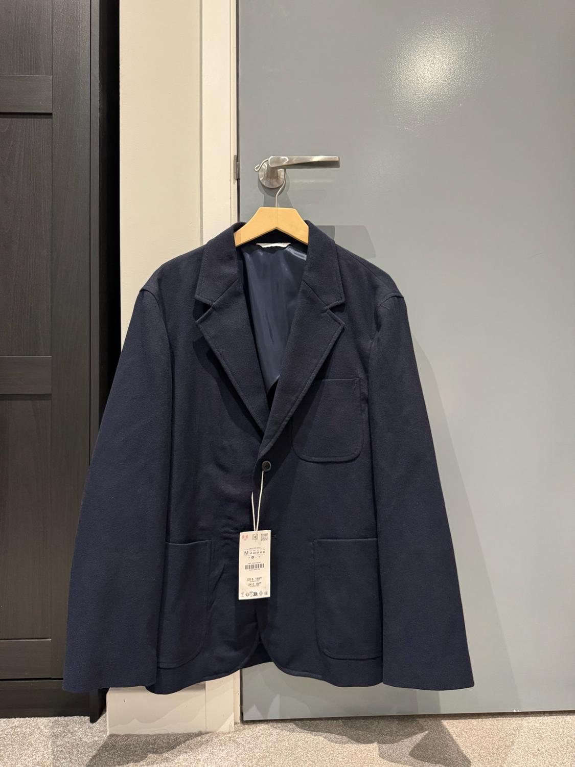 Navy blue jacket with pockets and a label, hanging from a wooden hanger on a grey door knob.