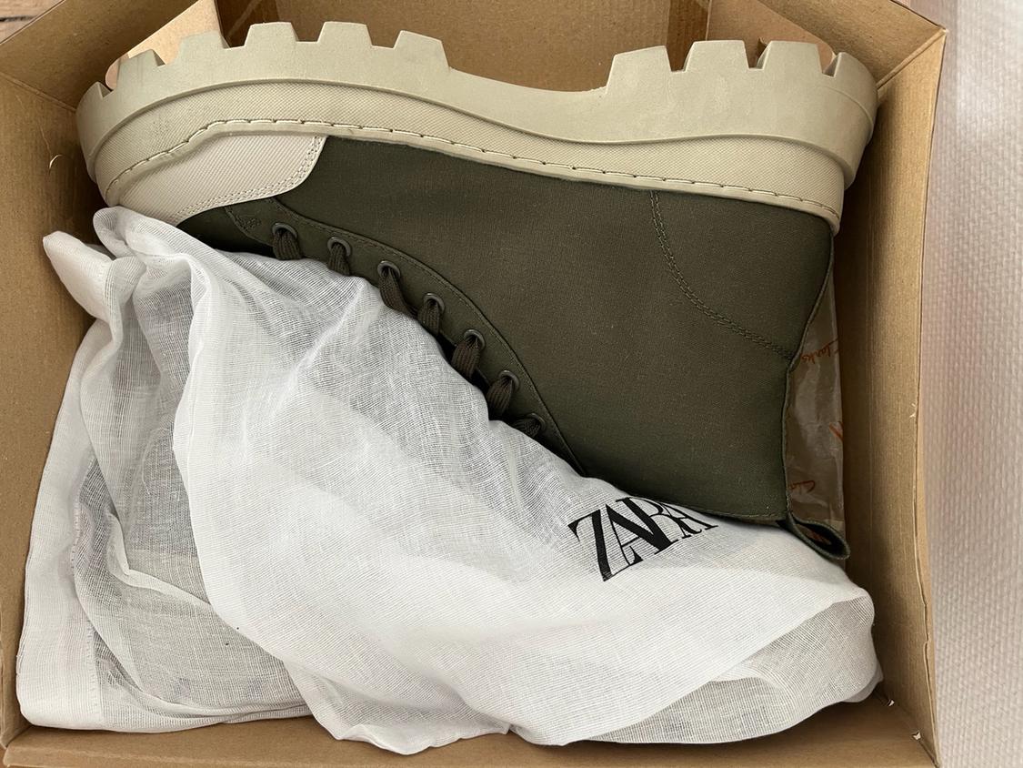 Olive green boot with a thick beige sole inside a cardboard box, on top of a white bag with the text ZARA.