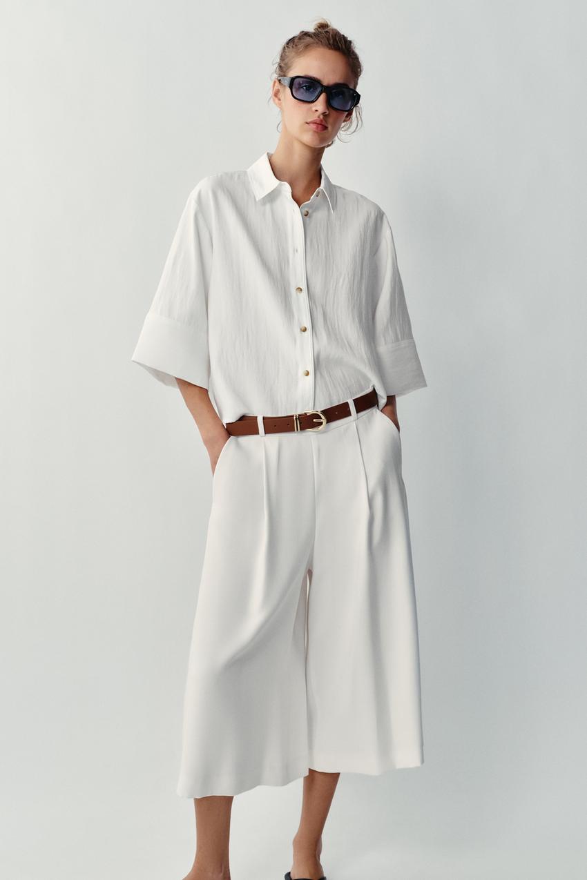 Conjunto de camisa blanca de manga corta, bermuda larga blanco roto con pinzas y bolsillos frontales, cinturón marrón y gafas de sol.