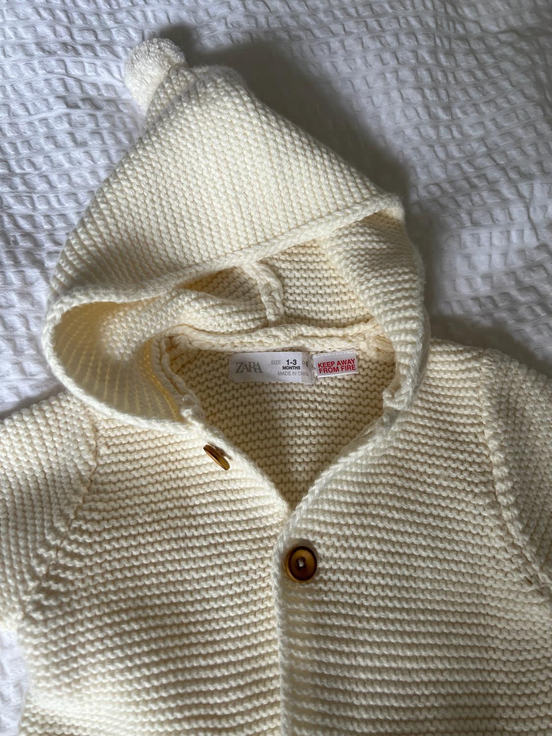 Cream knitted hooded cardigan over white fabric. Text: ZARA SIZE 1-3 MONTHS MADE IN CHINA KEEP AWAY FROM FIRE.