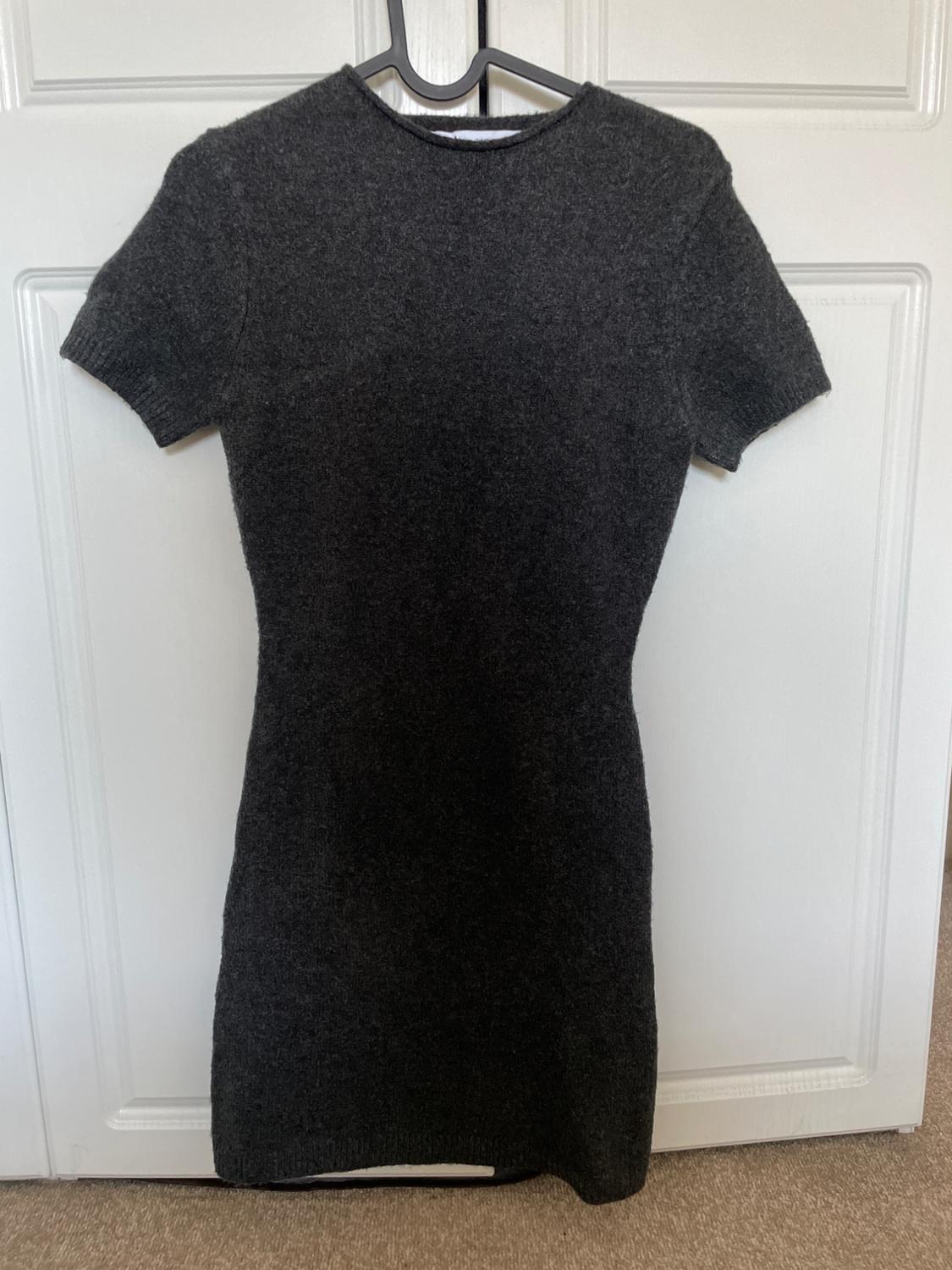 Dark grey knitted dress, short sleeves and round neck, hanging on a black hanger over a white door. Casual and relaxed atmosphere.