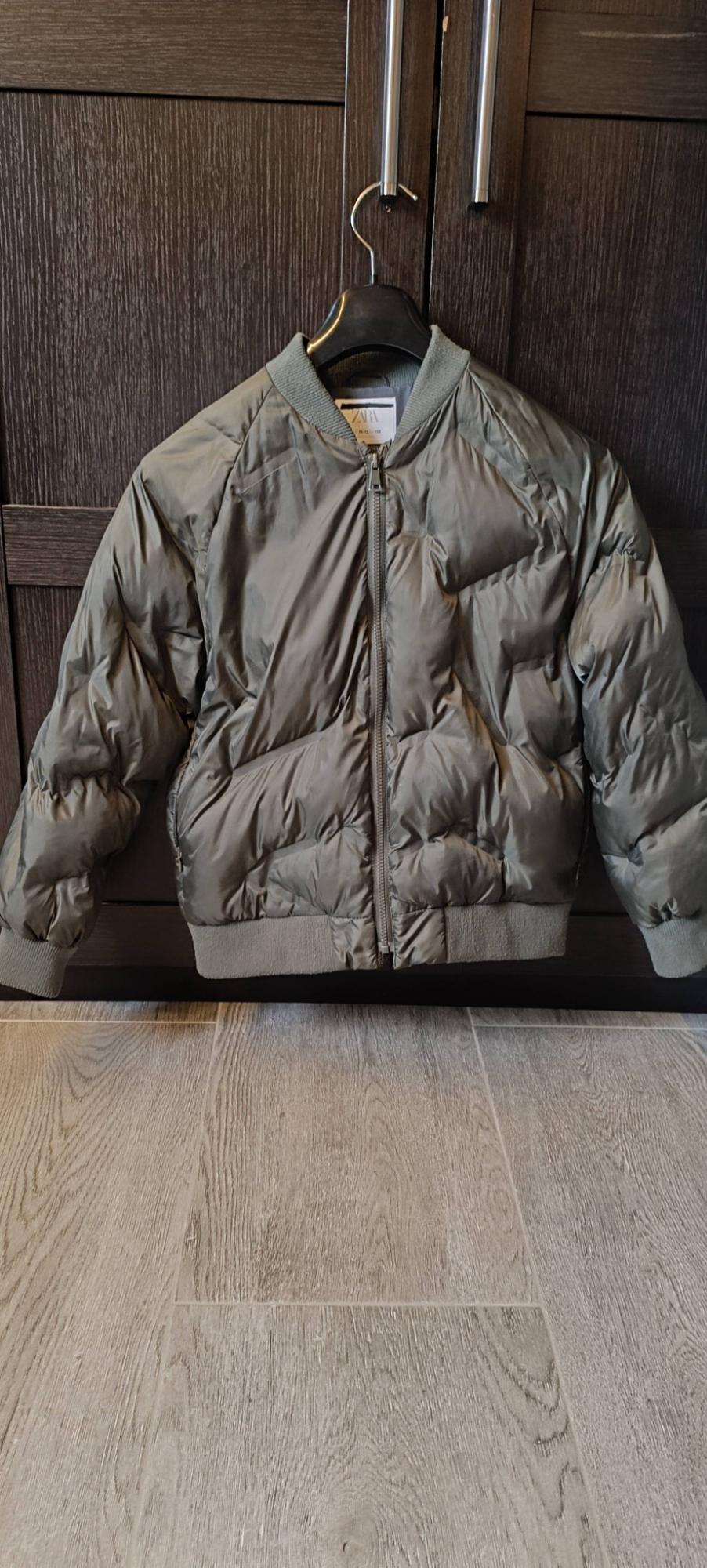 Olive green padded jacket with zip, hanging in a dark wood wardrobe over a grey floor. ZARA label inside.