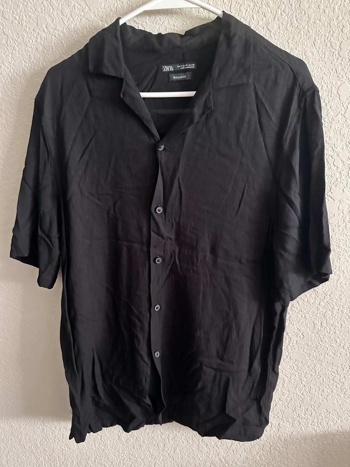 Short-sleeved black shirt hanging on a white coat hanger, with front buttons and open collar, against a light-coloured wall.