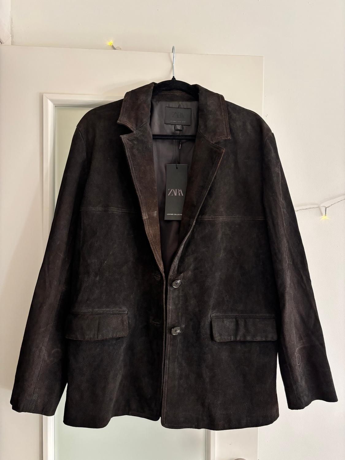 Black jacket hanging on a white door, with visible Zara tag. Classic style with lapels and front pockets.