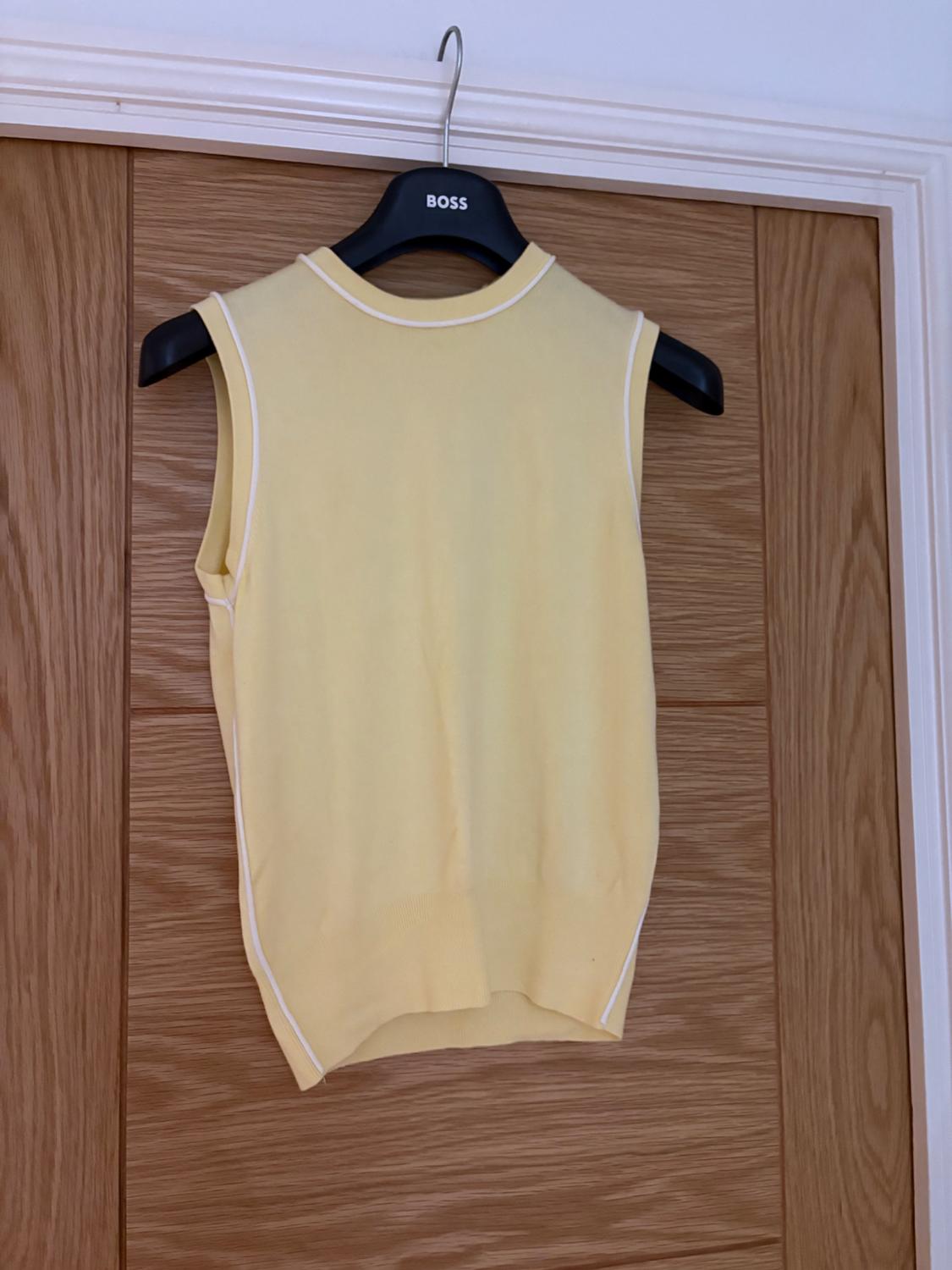 Yellow point sleeveless top with white trims, on a black hanger with the BOSS text, over a wooden door.
