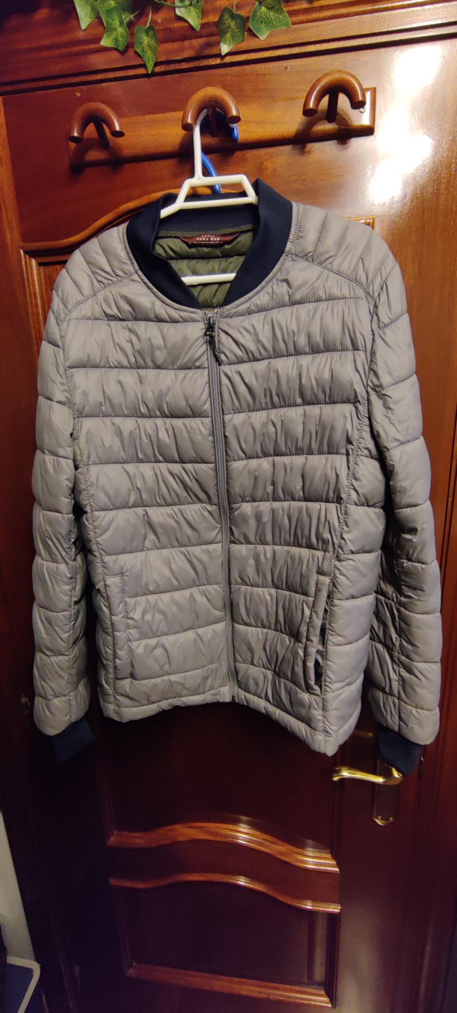 Grey quilted jacket with a dark collar, hanging on a hanger on a wooden door. Label text: ZARA MAN.