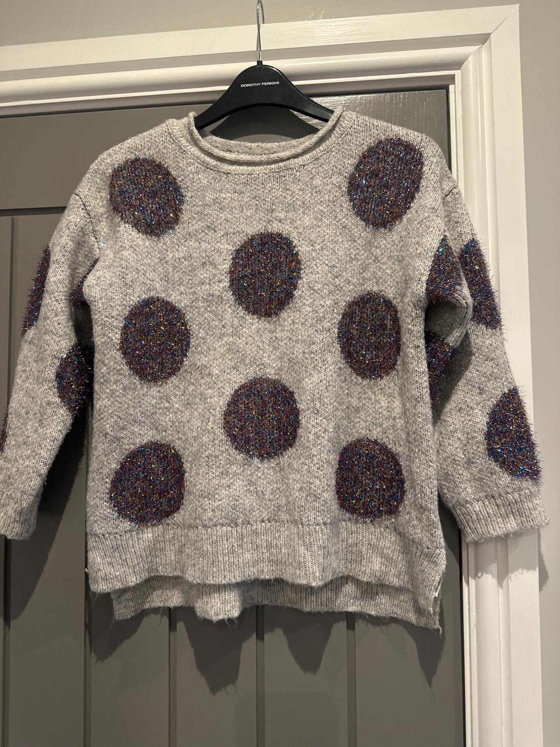 Grey jumper with large, sparkly polka dots in dark shades, hanging on a black coat hanger over a grey door.