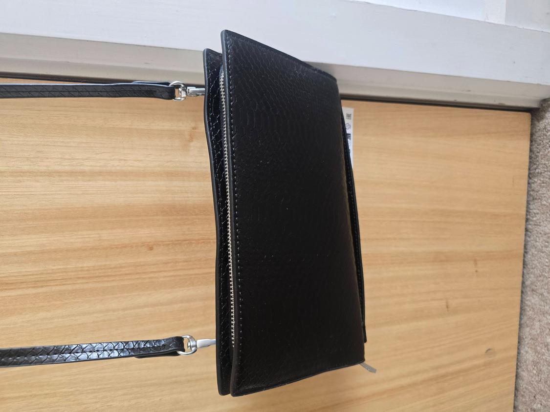 Black leather-look textured bag with a zip fastening, hanging from a strap on a light wooden surface.