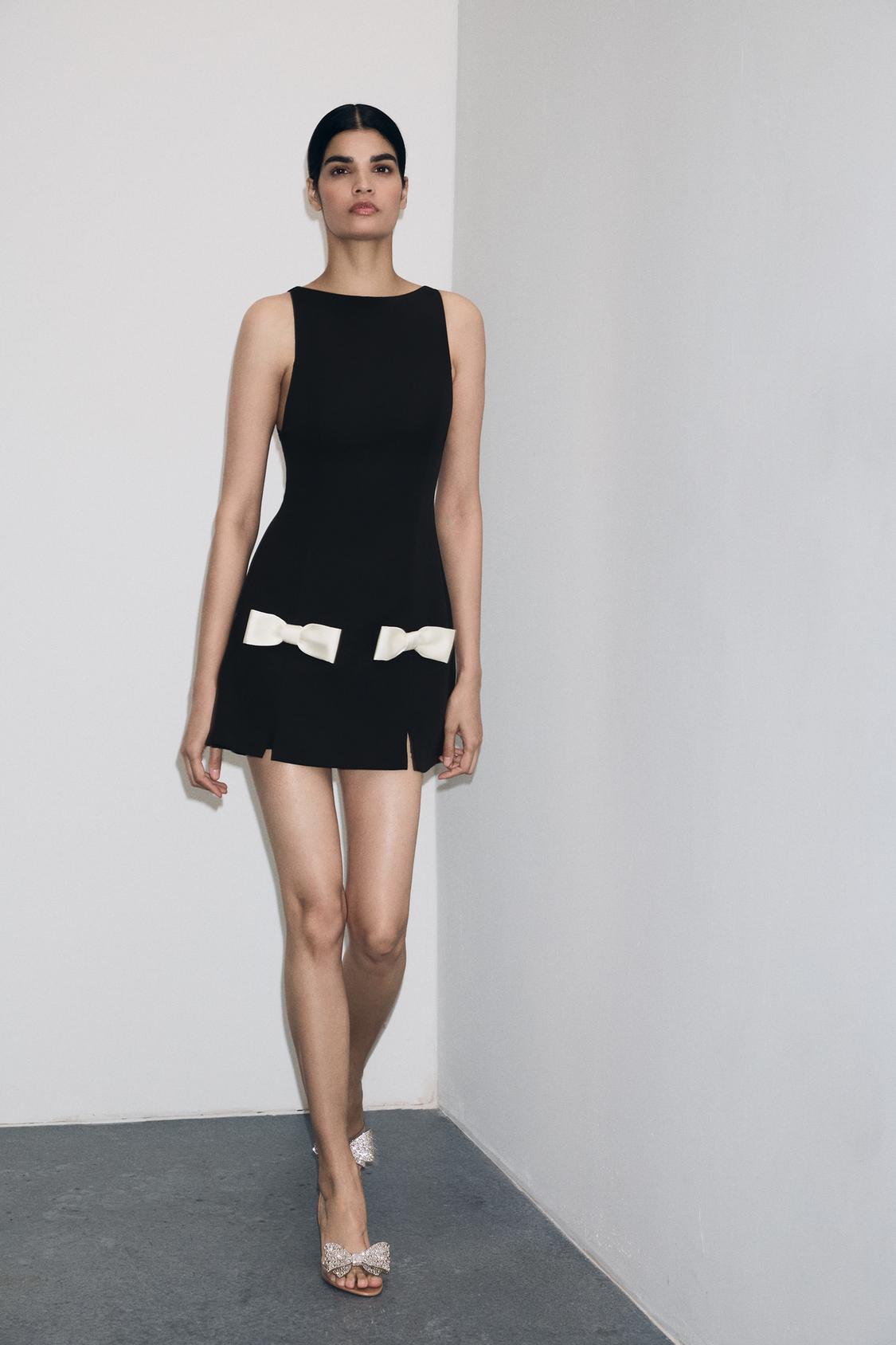 Komplet črne obleke brez rokavov z belimi mašnicami v pasu in sandali s sijočimi mašnicami.