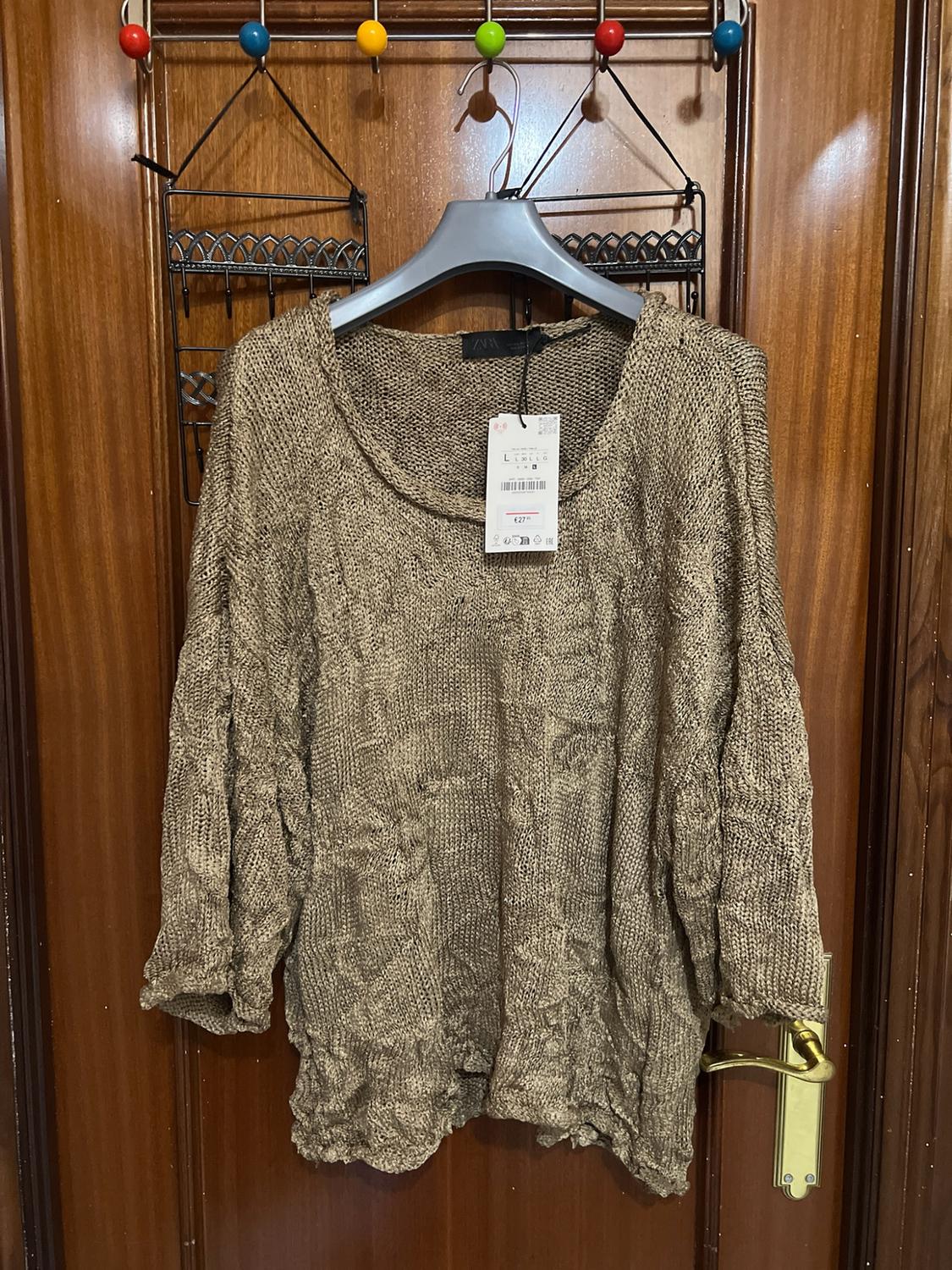 Gold knitted jumper with an openwork texture, three-quarter-length sleeves and a wide neckline. It is hanging on a coat hanger over a wooden door.