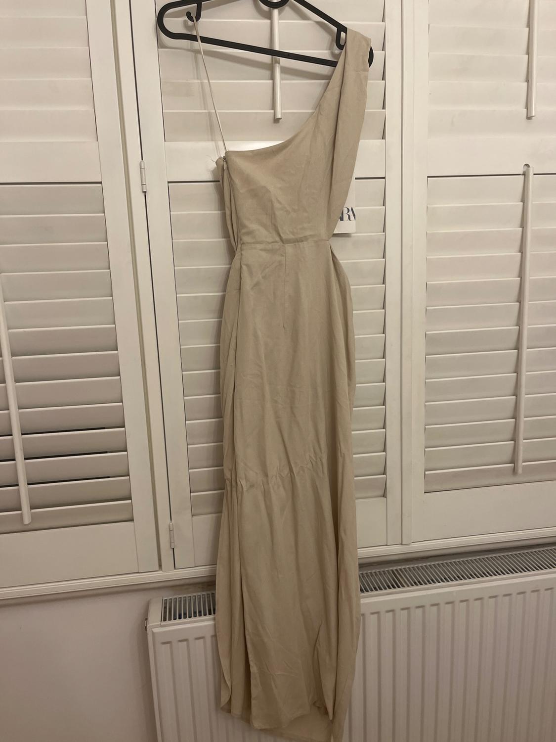 Beige, one-shoulder maxi dress hanging on a black coat hanger in front of white blinds and a white radiator.