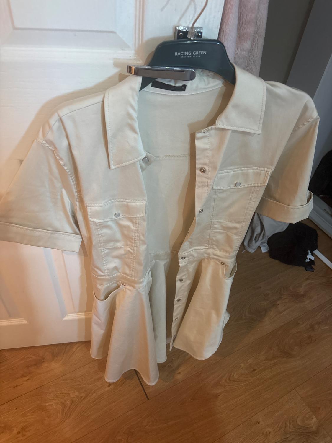 Beige shirt dress hanging on a white door. Black hanger with the text RACING GREEN BRITISH STYLE. Casual setting with wooden flooring.