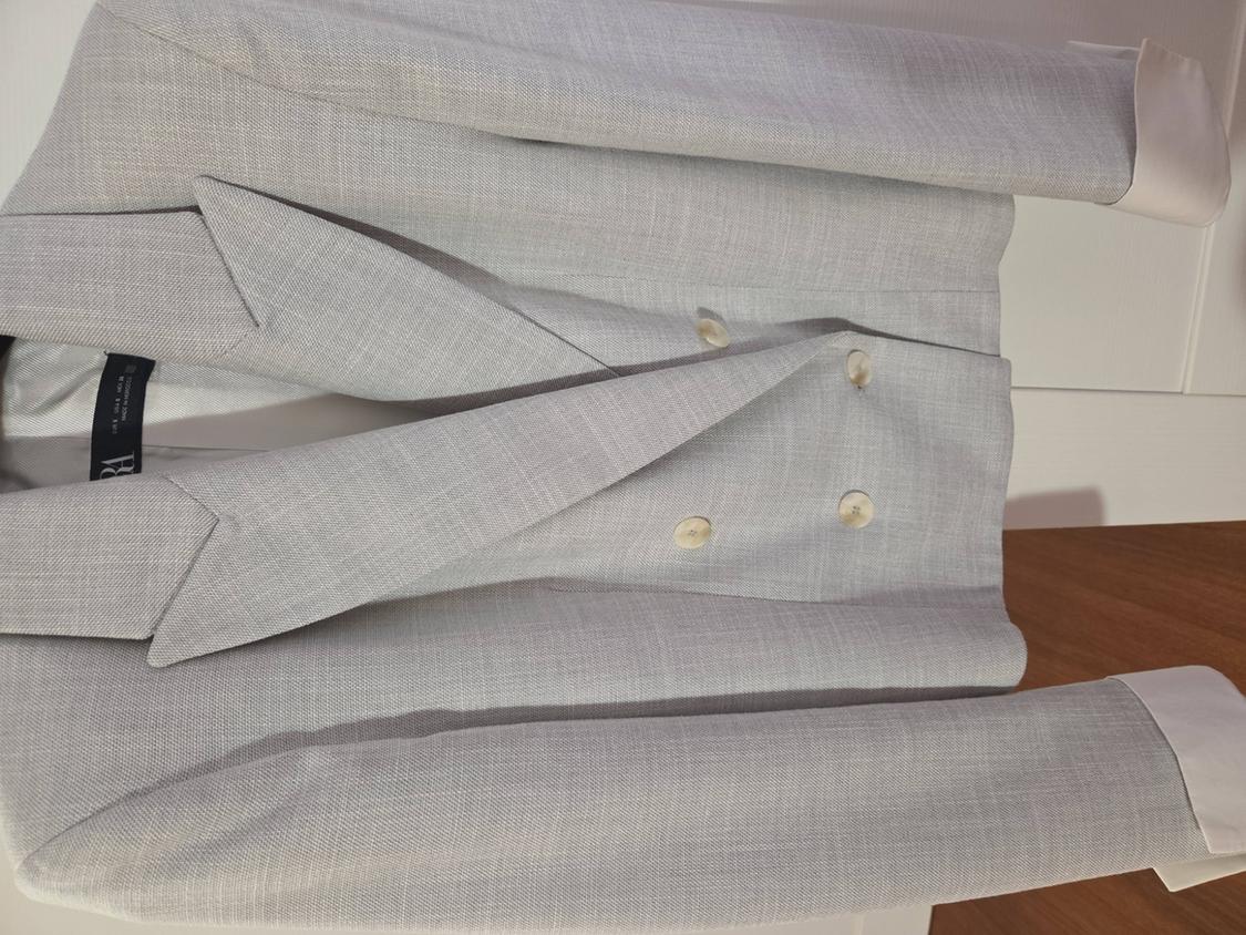 Light grey double-breasted jacket with beige buttons, hung on a wooden surface and white background.