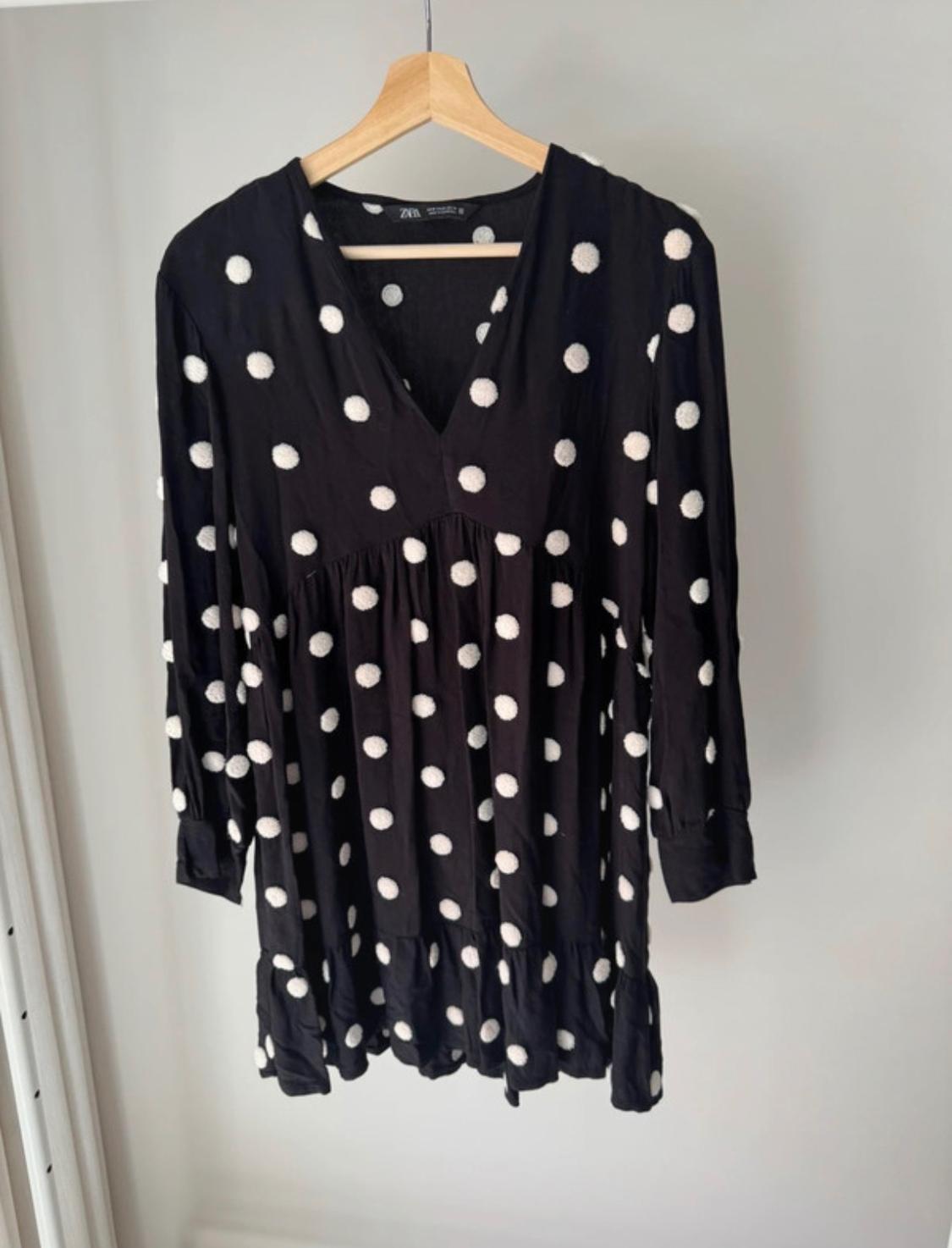 Black dress with white polka dots hanging on a wooden hanger. It has long sleeves and a V-neck. Light grey background.