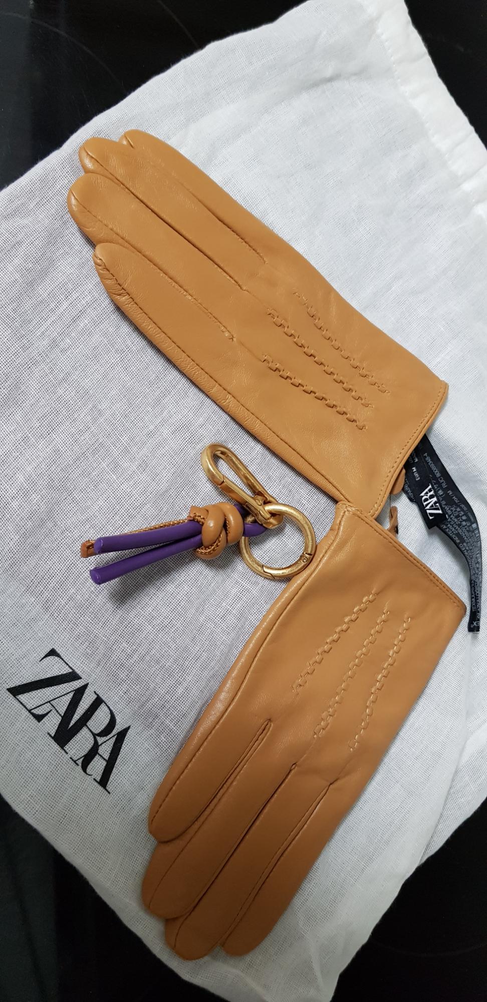 Brown gloves with decorative stitching and a gold keyring on a white bag with 'Zara' written on it.