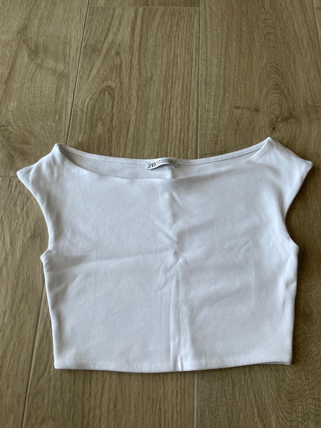 White short-sleeved top on a light wood floor. Visible neck label with "ZARA" text.