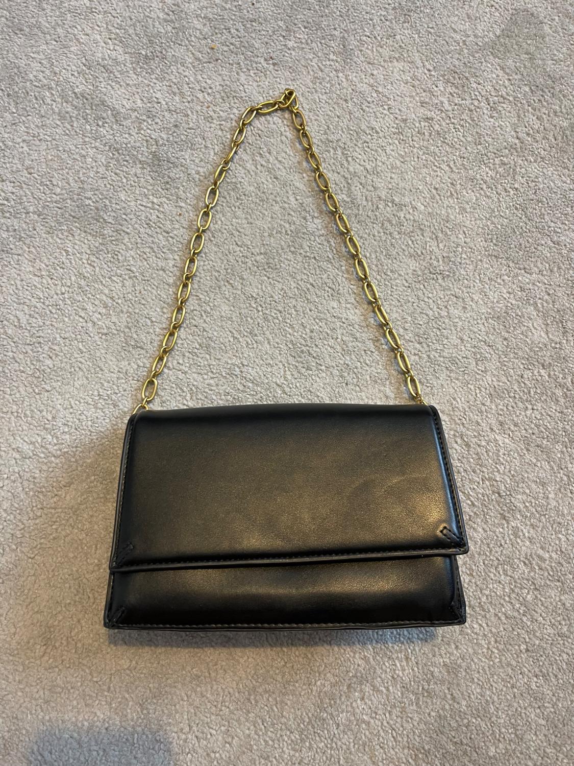 Black faux leather handbag with a gold chain on a light grey fabric background.