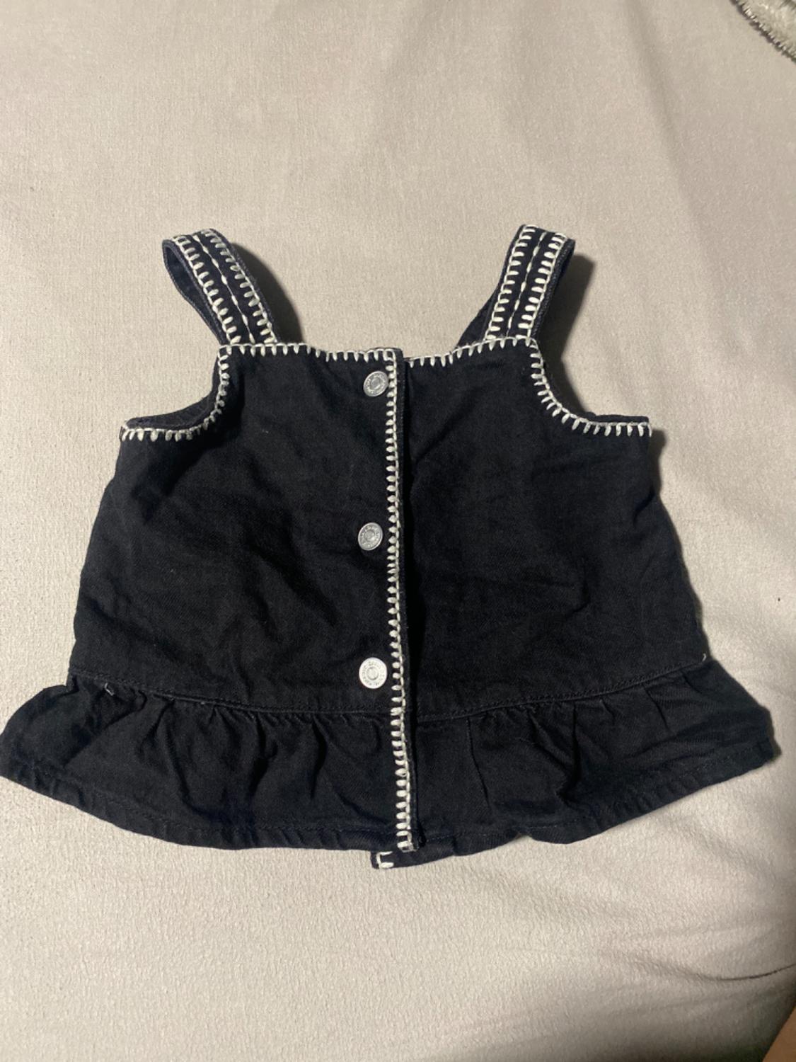 Black top with straps and silver buttons on a grey background. White stitching details on edges and straps.