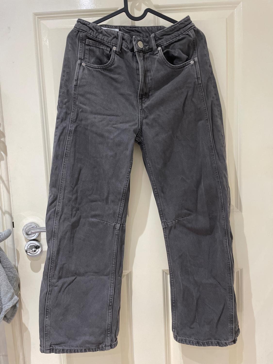 Dark grey jeans with front seams, hanging on a black hanger in front of a white door in a relaxed setting.