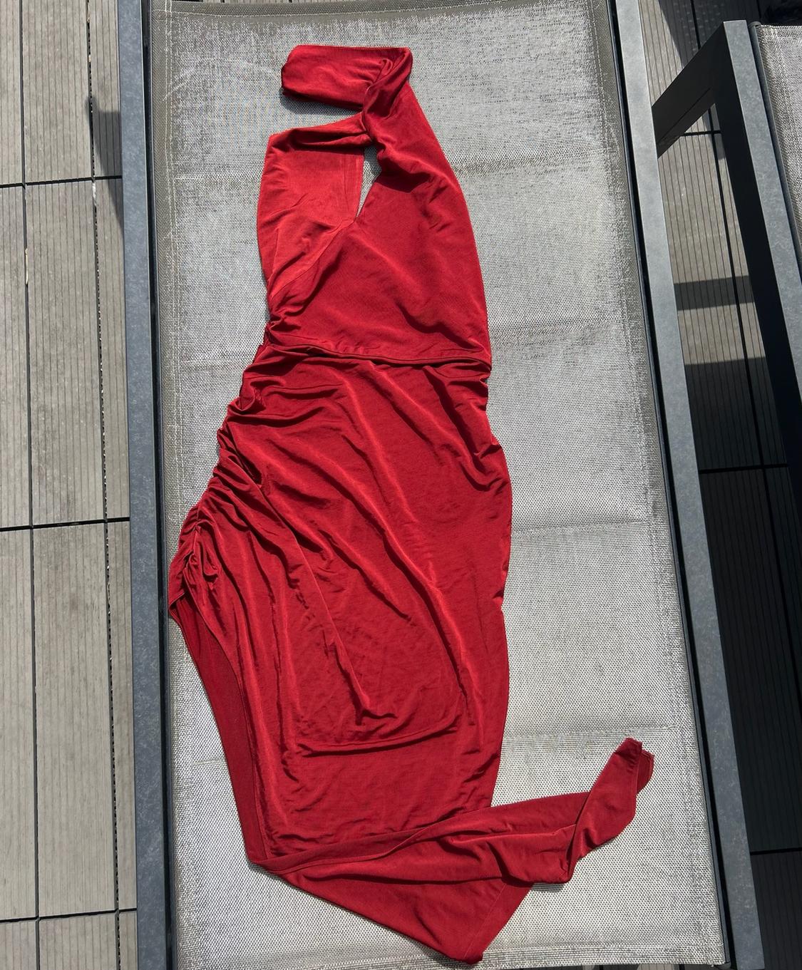 Long red dress with an asymmetric neckline and side ruching, laid out on a grey sun lounger on a sunny terrace.