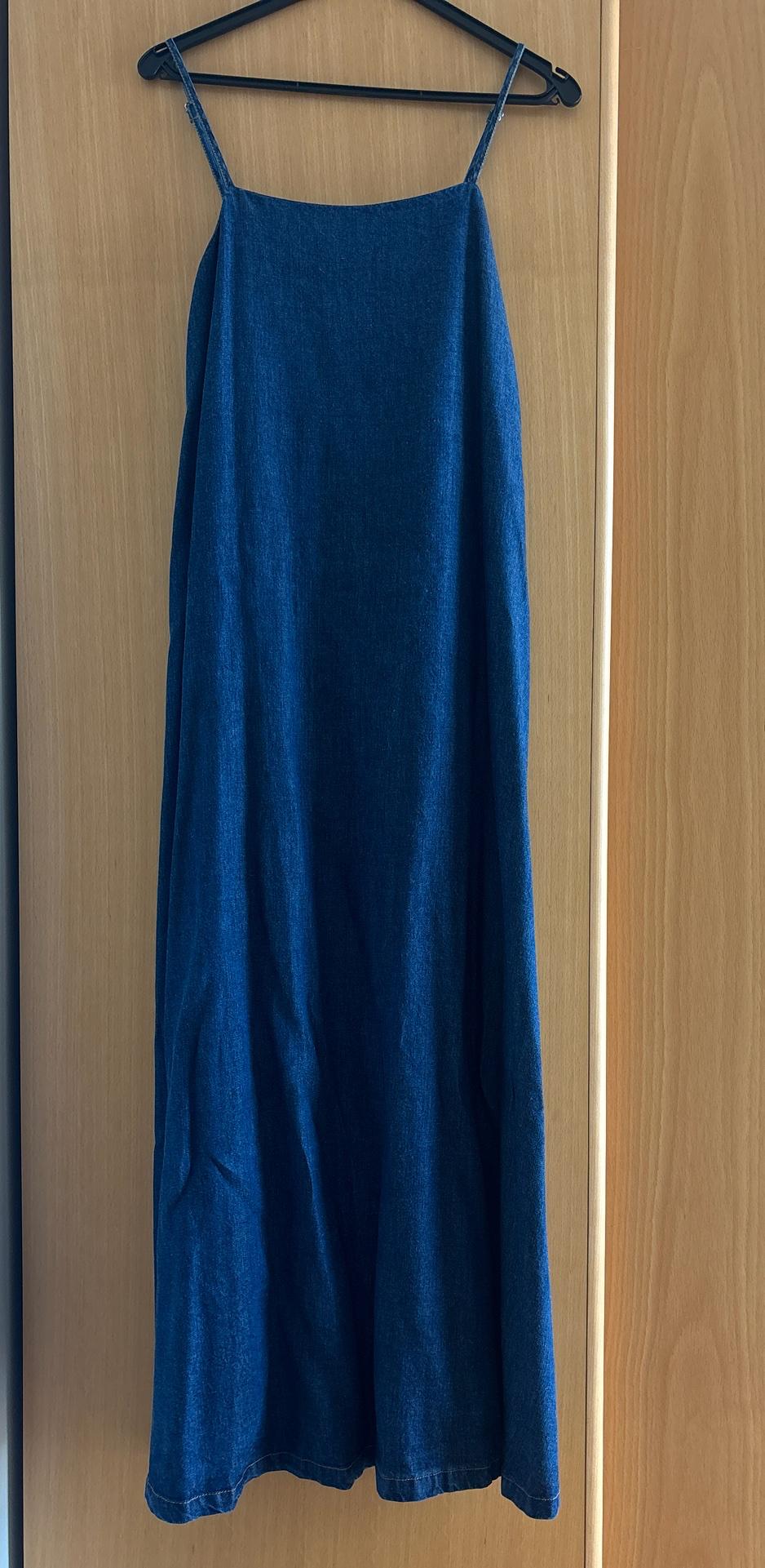 Dark blue denim maxi dress with thin straps, hanging on a black hanger in front of a wooden door.