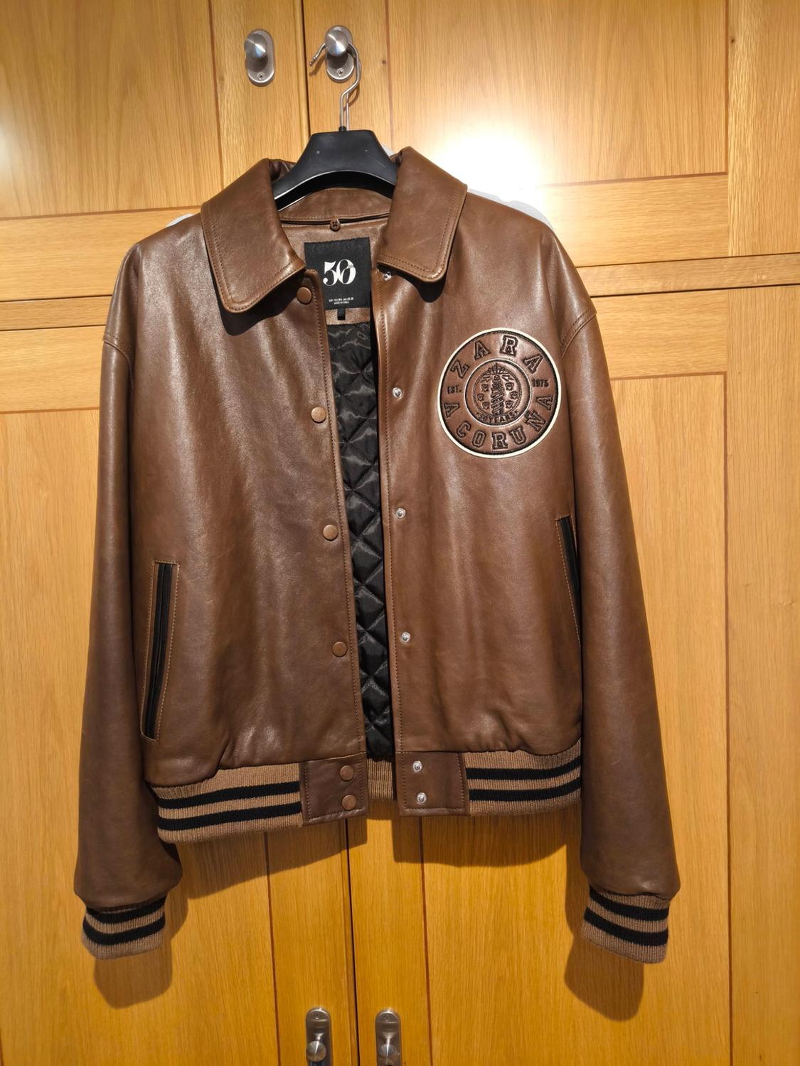 Brown jacket hanging in a wooden wardrobe, with a patch that says "ZARA A CORUÑA" and a "50" label. Black and beige details.