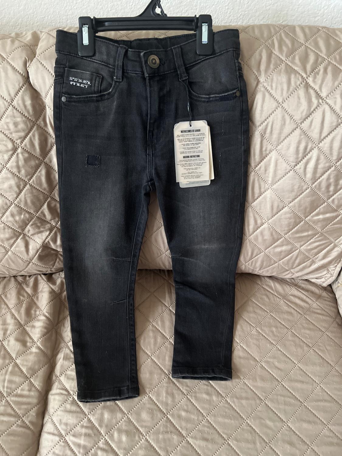 Black denim jeans hanging on a hanger over a beige upholstered sofa. Visible tag with washing instructions.