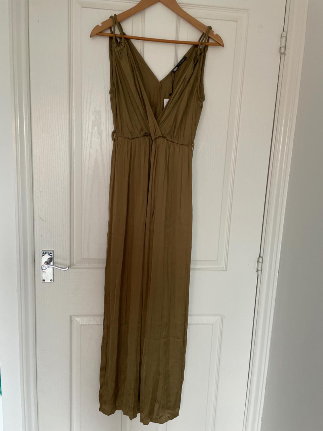 Olive green full-length jumpsuit with a V-neck, hanging on a wooden hanger against a white door. Relaxed, homely atmosphere. ZARA label.