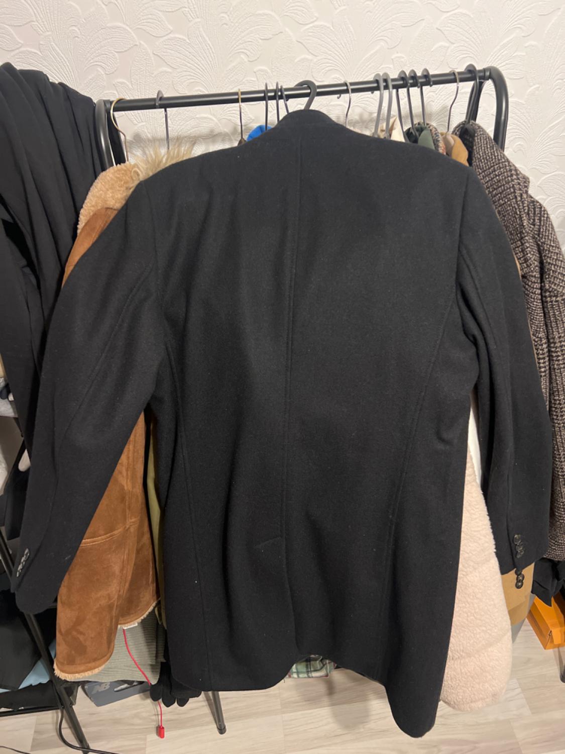 Black coat hanging on a clothes rail alongside other garments of different colours and textures. Background with a floral design wall.