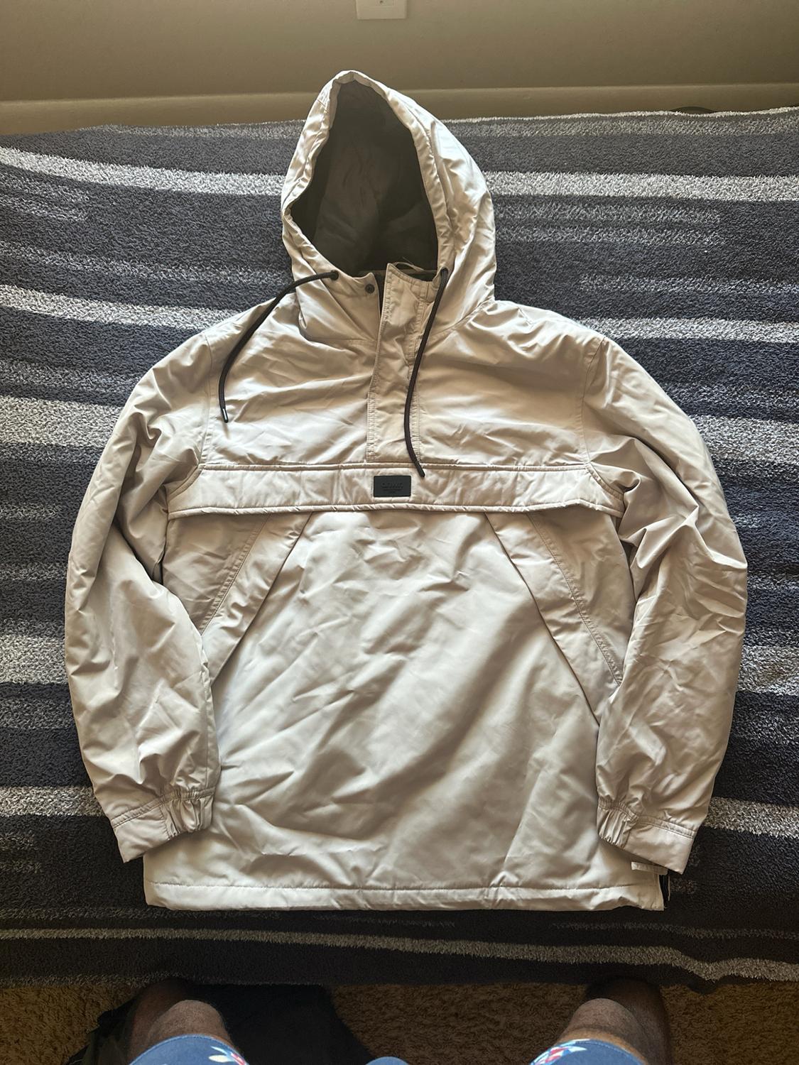 Beige kangaroo-style jacket with a hood and black drawstrings, spread over a gray-and-white striped blanket.