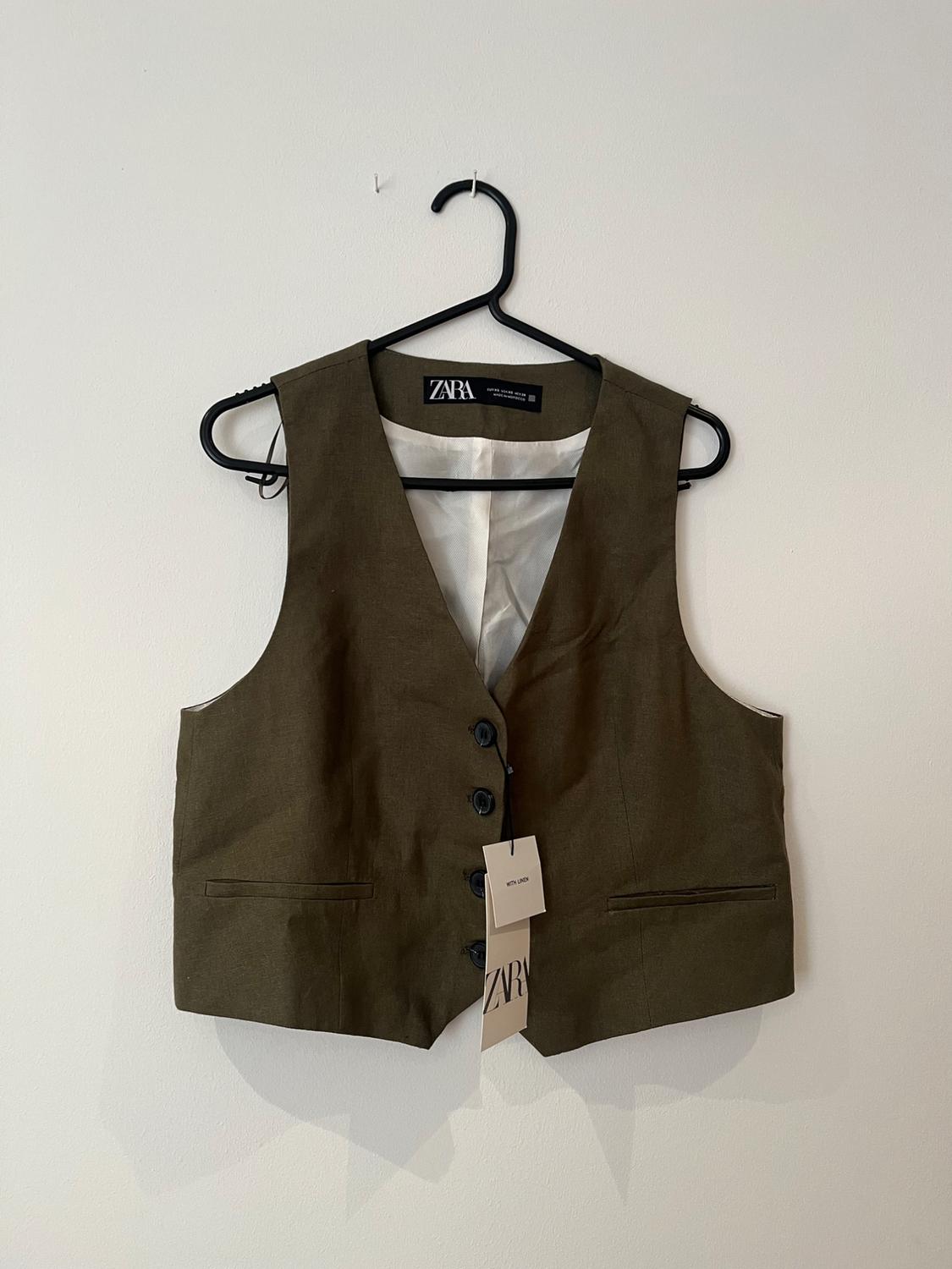 Brown waistcoat hanging on a black hanger against a white wall. Visible tag with the text "ZARA".
