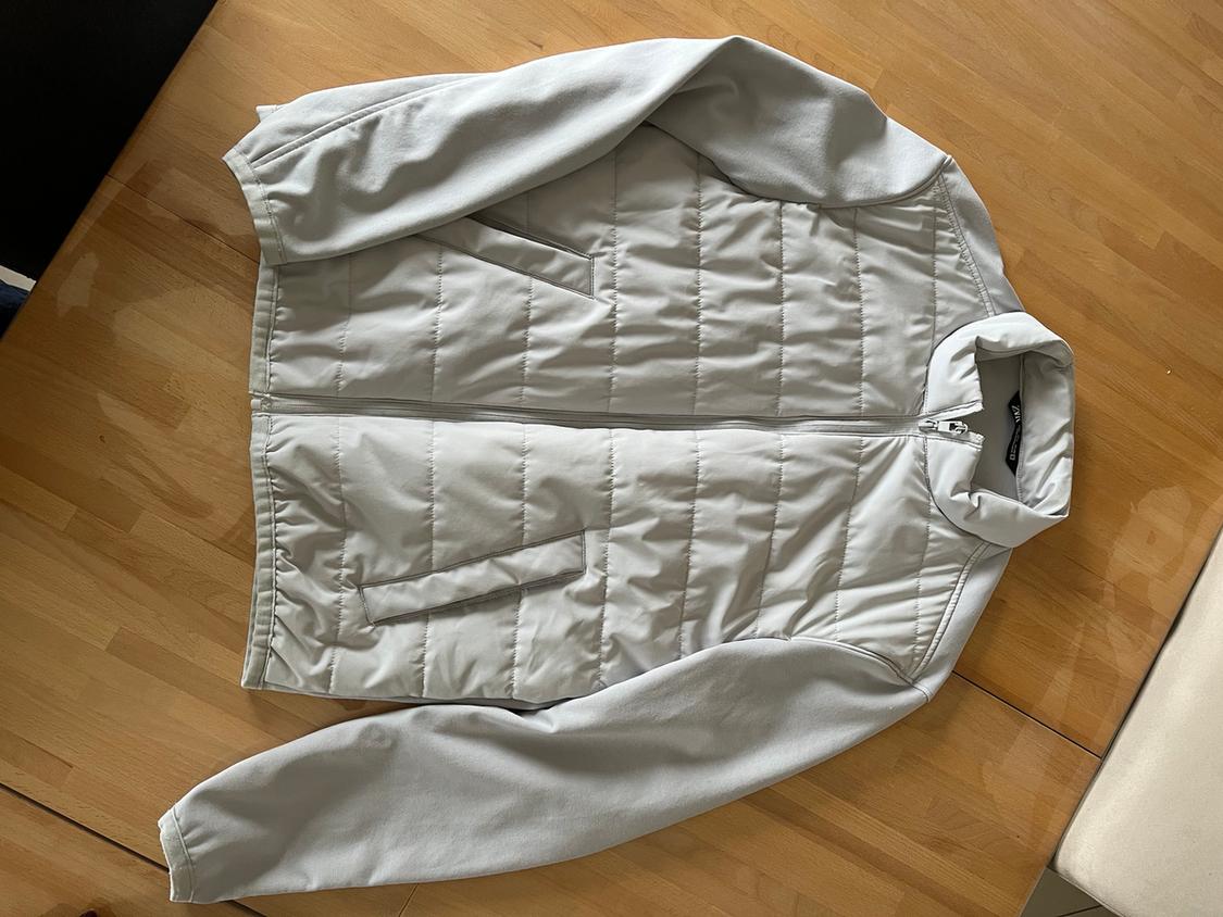 Light grey padded jacket with zip and pockets, on a wooden table. Plain sleeves and high collar.