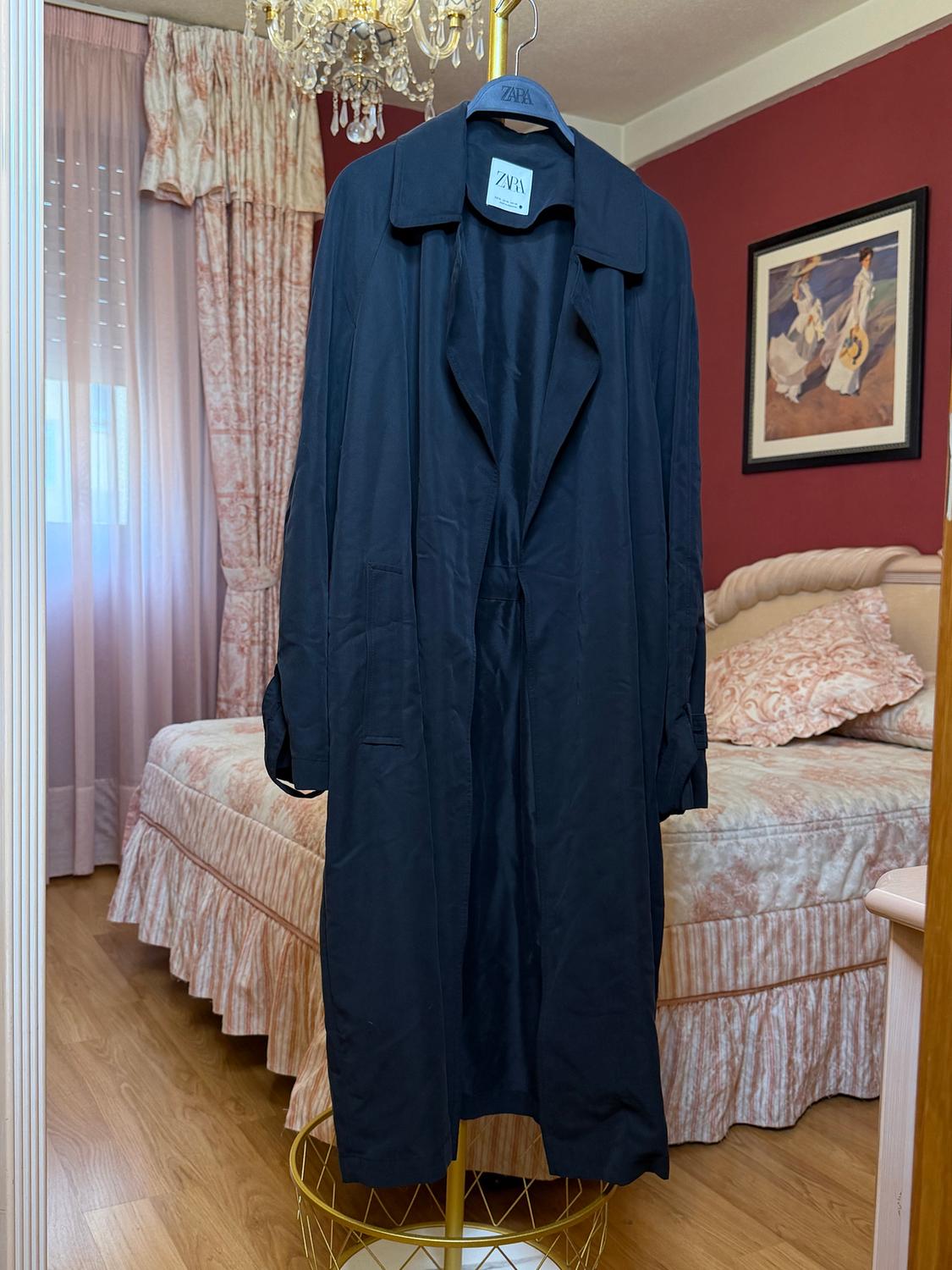 Black coat hanging on a Zara hanger, in a bedroom with pink curtains, a bed with a patterned bedspread, and a chandelier.
