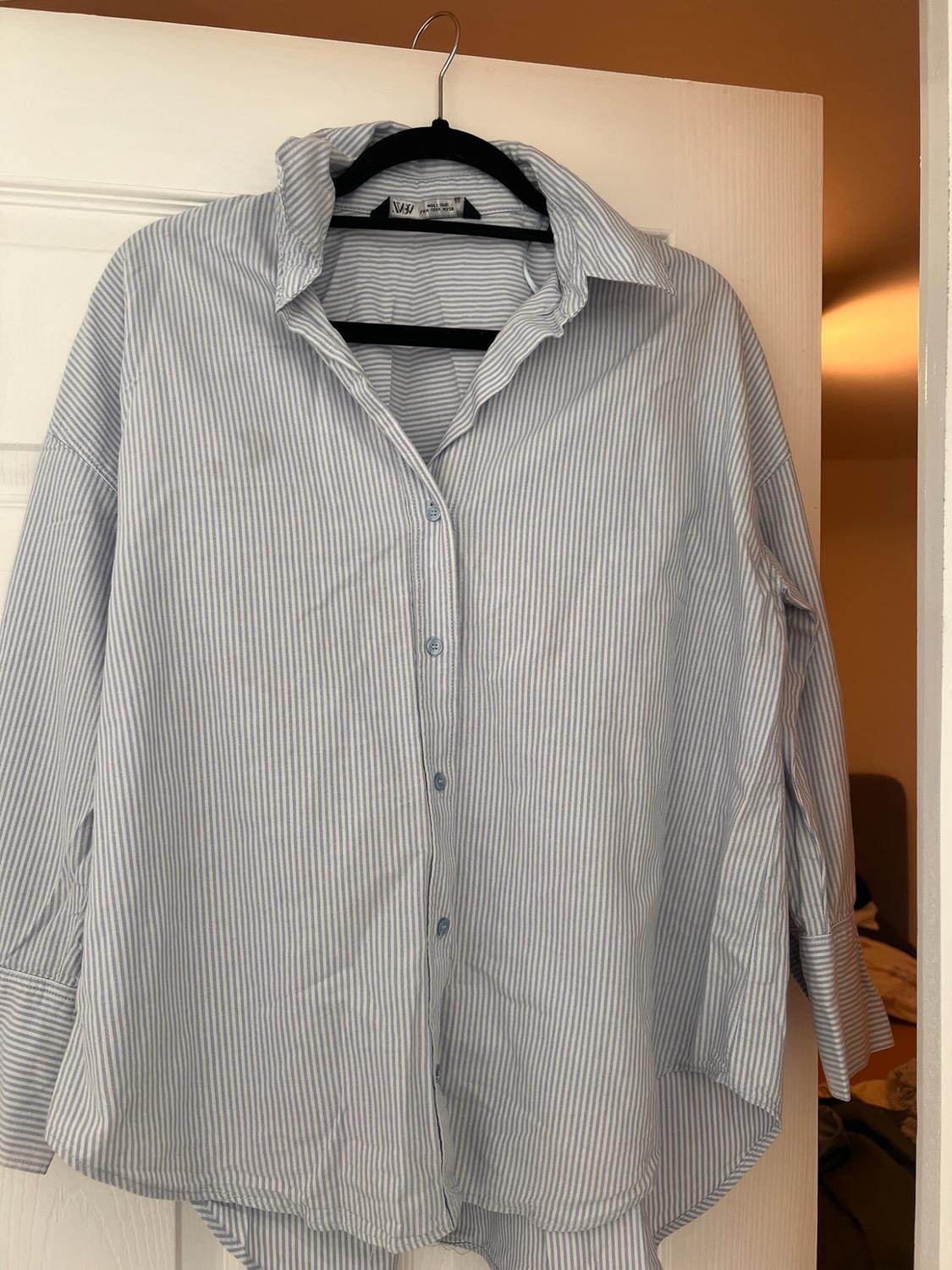 Loose blue and white striped shirt hanging on a black hanger against a white door. ZARA text on the label. Casual style.