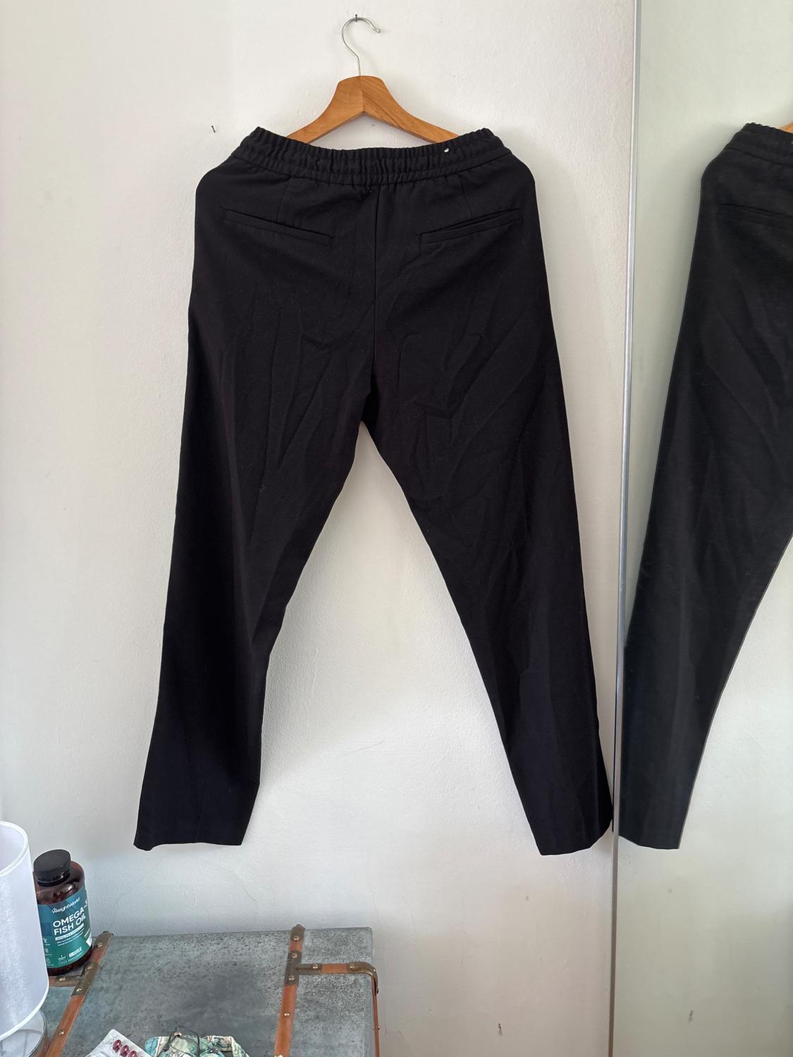 Black trousers with an elasticated waist hanging on a white wall next to a mirror. Below is a trunk and a tub that says OMEGA FISH OIL.